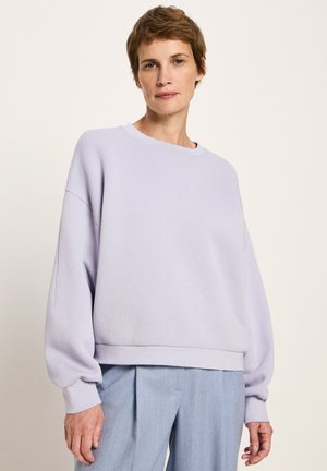 Frau mit kurzem braunem Haar, die einen lockeren, helllila Pullover und eine hellblaue, plissierte Hose trägt, steht vor einem schlichten Hintergrund.