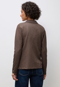 Camicia marrone a maniche lunghe con colletto, realizzata in tessuto morbido, caratterizzata da una texture liscia e un fit rilassato, abbinata a jeans blu.
