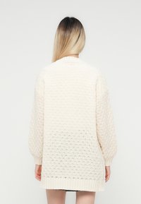 Femme avec de longs cheveux ombrés portant un pull en maille de couleur crème, vue de dos sur un fond uni clair.