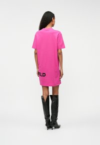 Vestido t-shirt oversized rosa com mangas curtas, decote redondo e um detalhe em texto preto com logo nas costas, combinado com botas pretas altas.