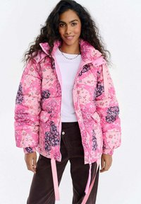 Różowa pikowana kurtka z kwiatowymi wzorami, puchowy fason, wysoki kołnierz i regulowane sznurki. Zestawiona z białą koszulą i brązowymi spodniami.