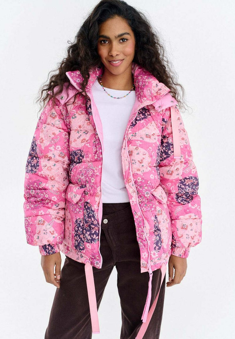 Różowa pikowana kurtka z kwiatowymi wzorami, puchowy fason, wysoki kołnierz i regulowane sznurki. Zestawiona z białą koszulą i brązowymi spodniami.