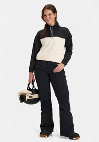 Melns un krēmkrāsas džemperis ar augstu apkakli, melnas slēpošanas bikses un melna ķivere ar krēmkrāsas polsterējumu, turēta rokā. Mūsdienīgs, sportisks dizains.