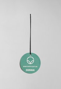 Pyöreä vihreä hangtag mustalla narulla, jossa on valkoista tekstiä "GOOD EARTH COTTON" ja logo, joka muistuttaa lehden ääriviivaa, Napapijriltä.