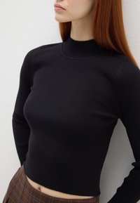Pull en maille côtelée noir avec un col montant et des manches longues, présentant un design court, porté avec une jupe vichy marron.