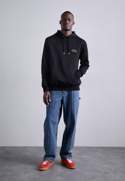 Sudadera negra con capucha, bolsillo frontal y detalles en dorado, combinada con unos jeans de rayas azul claro y zapatillas rojas con una suela marrón.