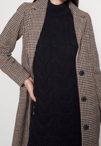 Cappotto houndstooth marrone e nero con grandi bottoni, abbinato a un maglione nero in lavorazione a trecce. Il cappotto ha un colletto a rever.