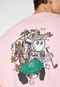 Rosa T-skjorte med et tegneserie-design av en hund og en golfball som kjører en grønn vogn, med golfkøller og golfballer i bakgrunnen.
