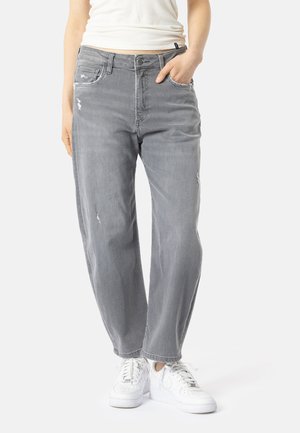 Persona con jeans grises desgastados y zapatillas blancas, de pie con una mano en el bolsillo contra un fondo liso.