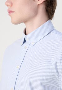 Chemise à rayures bleu clair et blanches avec un col boutonné. Les caractéristiques incluent des fermetures à boutons et un petit logo brodé sur la poitrine.