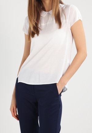 Femme portant un chemisier blanc à manches courtes avec col et un pantalon bleu marine, debout avec une main dans la poche, sur un fond uni.
