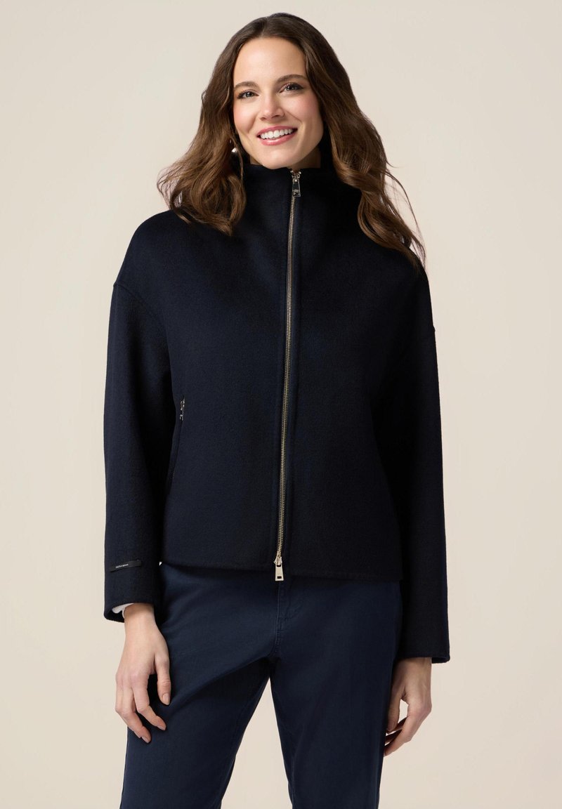 Chaqueta azul marino con cremallera, hecha de material suave, que cuenta con un alto cuello, mangas anchas y bolsillos laterales con detalles de hardware plateado.