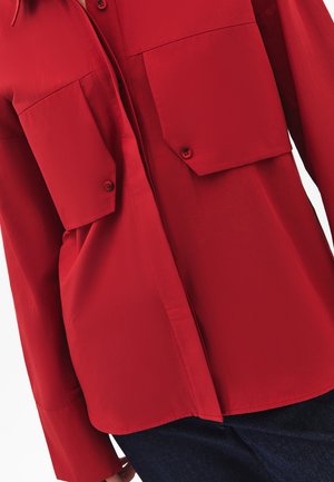 Chemise rouge avec deux grandes poches poitrine, manches longues et ourlet droit. Texture lisse avec détails en boutons.
