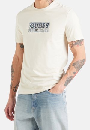 Mężczyzna w białej koszulce Guess z niebieskim logo "GUESS EST. 1981" i jasnoniebieskich dżinsach, pokazujący tatuaże na obu ramionach.