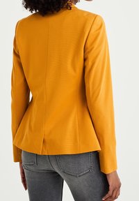 Senapsgul skräddarsydd kavaj med strukturerade axlar och en peplum-siluett, tillverkad av ett jämnt, texturerat tyg.