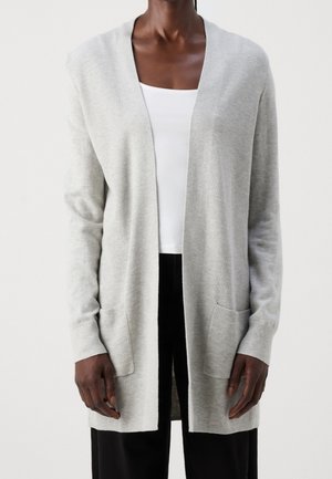 Person iført en lysegrå cardigan med åben front, lange ærmer og forlommer over en hvid top og sorte bukser.