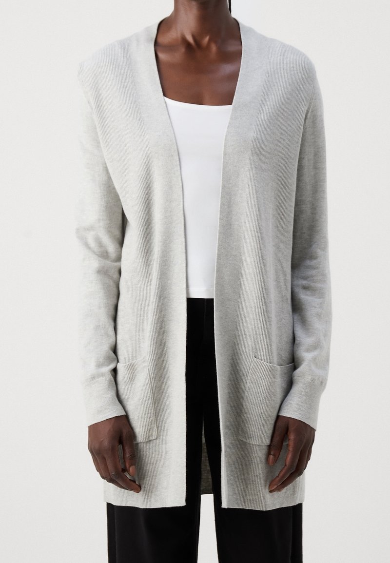 Personne portant un cardigan gris clair ouvert à l'avant avec des manches longues et des poches devant, par-dessus un haut blanc et un pantalon noir.