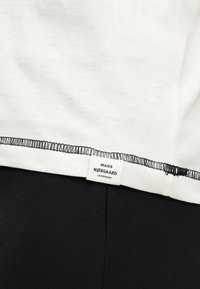 Κοντινή λήψη της γωνίας ενός λευκού ενδύματος με μαύρο γαζί overlock και μια μικρή ετικέτα που γράφει «MADS NØRGAARD COPENHAGEN» πάνω σε μαύρο φόντο υφάσματος.