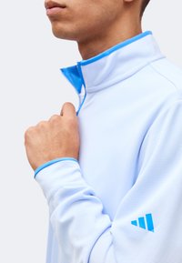 Άνδρας τραβάει το φερμουάρ ενός γαλάζιου αθλητικού μπουφάν με μπλε περιθώριο στο γιακά και λογότυπο Adidas στο μανίκι.