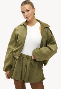 Veste vert olive à coupe décontractée avec fermeture éclair à l'avant et manches larges, associée à une jupe à cordon de serrage assortie. Débardeur blanc à col rond en dessous.