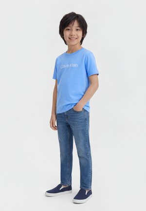 Sötét hajú fiú világoskék Calvin Klein pólót, kék farmert és sötétkék slip-on cipőt visel, egyik kezét a zsebében tartva áll.