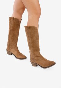 Botas de ante color beige hasta la rodilla con una forma estructurada, puntera redondeada y un tacón apilado, que cuentan con acentos de costura y pestañas para ayudar a ponérselas.