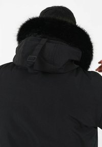 Svart huva jacka med en lurvlig krage, med en matt yta och justerbara remmar på huvan. Endast bakvy visas.