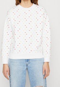 Felpa bianca con piccoli motivi rettangolari multicolori; presenta un colletto rotondo, polsini a costine e una vestibilità rilassata. Abbinata a jeans strappati.