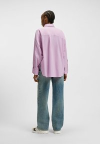 Camicia oversized di un viola chiaro con maniche lunghe, dettaglio della spalla sul retro, abbinata a jeans a gamba larga blu sbiaditi e sneakers bianche.