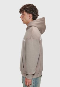 Junger Mann mit lockigem Haar, der einen taupefarbenen Hoodie mit Fronttasche und hellblauen Jeans trägt, im Profil vor einem schlichten Hintergrund. Expansion