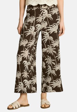 Brązowe spodnie o szerokich nogawkach z beżowym nadrukiem palm, noszone z beżowymi sandałami na płaskiej podeszwie z odkrytymi palcami oraz białą szydełkowaną bluzką.