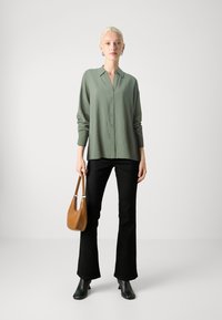 Camisa de botão verde-claro com mangas longas, combinada com calças pretas à boca de sino, usando botas pretas de salto alto e segurando uma bolsa castanha.