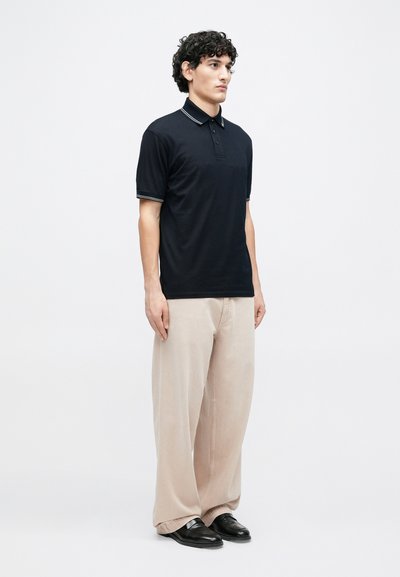 Μπλε ναυτικό πόλο μπλουζάκι με γιακά και τρία κουμπιά, με λευκές ριγέ λεπτομέρειες στα μανίκια. Συνδυασμένο με μπεζ παντελόνι φαρδιάς γραμμής και μαύρα παπούτσια.
