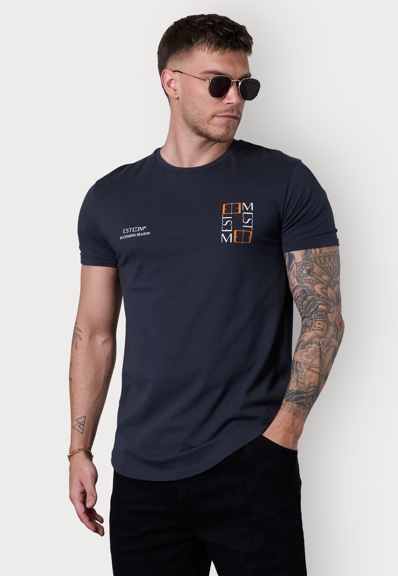 T-shirt bleu marine en coton, avec un col rond, des manches courtes et un logo imprimé en orange et blanc sur la zone de la poitrine gauche.