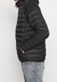 Doudoune noire avec une fermeture éclair à l'avant, des coutures horizontales et deux poches latérales. Elle dispose d'une capuche et est fabriquée dans un matériau brillant et lisse.