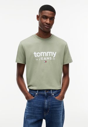 Mężczyzna w jasnozielonej koszulce Tommy Jeans i niebieskich dżinsach, stojący z rękami w kieszeniach na jednolitym białym tle.