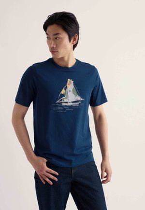 Homme portant un t-shirt bleu marine avec un graphisme coloré de voilier, debout, une main dans la poche et regardant sur le côté.