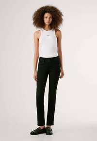 Vaqueros negros ajustados con diseño de cinco bolsillos, cierre de botón y bragueta frontal, combinados con una camiseta blanca sin mangas con un logo.