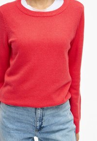 Personne portant un pull à manches longues corail vif par-dessus une chemise blanche, associé à un jean en denim bleu clair, sur un fond uni.