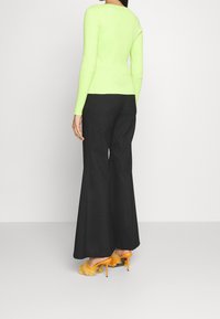 Kvinna i en neongrön ribbad långärmad tröja, svarta vida byxor och orange högklackade tofflor med fluffiga pälsdetaljer, stående.