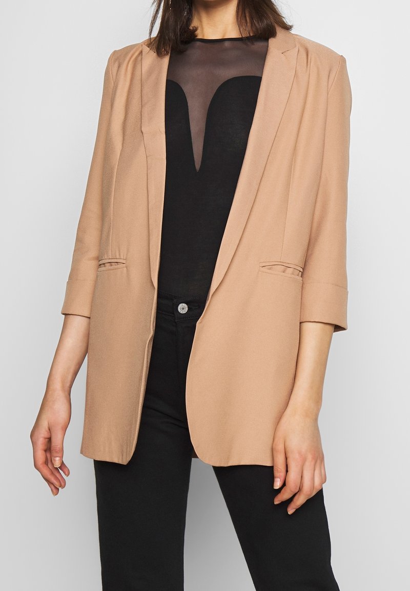 Femme portant un blazer beige sur un haut noir à encolure transparente et un pantalon noir, debout devant un fond clair uni.