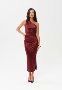 Bordeaux satijnen jurk met één schouderontwerp, plooidetail bij de taille en een hoge split. Gecombineerd met puntige, donkere hakken.