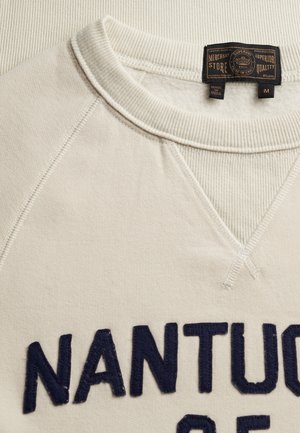 Primer plano del escote de una sudadera beige con cuello acanalado, etiqueta negra y la palabra "NANTU" visible en letras bordadas de color azul oscuro.