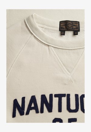 Primer plano del escote de una sudadera beige con cuello acanalado, etiqueta negra y la palabra "NANTU" visible en letras bordadas de color azul oscuro.