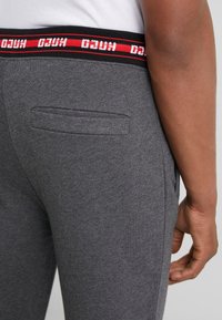 Graue Jogginghosen mit einem elastischen Bund in Schwarz und Rot, auf dem das "HUGO"-Logo prangt. Sie verfügen über eine einzelne Gesäßtasche und sind aus weichem, strukturiertem Stoff gefertigt.