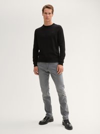 Hombre de pie con un suéter negro de manga larga, jeans grises y zapatos de cuero negros frente a un fondo claro y liso.
