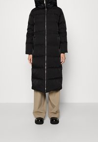 Schwarze Puffy-Jacke mit Reißverschluss vorne, Kapuze und horizontalen Nähten. Glatte Textur, knielanges Design, kombiniert mit beigen Hosen und schwarzen Schuhen.