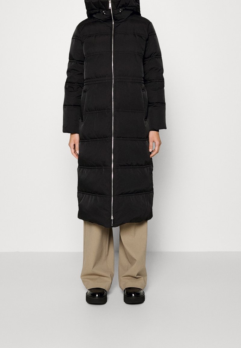 Schwarze Puffy-Jacke mit Reißverschluss vorne, Kapuze und horizontalen Nähten. Glatte Textur, knielanges Design, kombiniert mit beigen Hosen und schwarzen Schuhen.