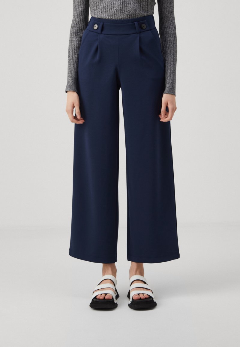 Pantalon large bleu marine avec plis à l'avant, deux boutons à la taille, associé à des sandales noires avec des lanières blanches.