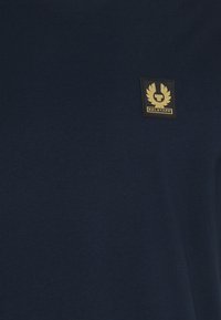 Tamsiai mėlynas marškinėliai, pagaminti iš glotnaus audinio, su juodu ir auksiniu logotipo įsiuvu ir lauro detale kairiajame krūtinės plote.